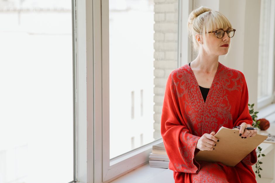 An elegant woman in glasses and red robe holding a clipboard, seated by a window.